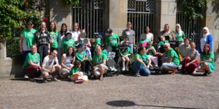 Gruppenbild Talker Camp 2026 Eine größere Gruppe von Menschen posiert im Freien für ein Gruppenfoto vor einem Gebäude mit steinernen Bögen, Metallgittern und Kletterpflanzen. Viele tragen grüne T‑Shirts mit einem Aufdruck. Einige Personen sitzen oder knien vorne, andere stehen dahinter. Mehrere Teilnehmende nutzen Rollstühle oder andere Hilfsmittel. Der Platz vor ihnen ist mit Kopfsteinpflaster bedeckt, und es ist sonniges Wetter.